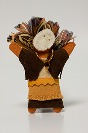 a small doll-like fetish with a headdress of feathers, beaded necklace, mask and dress made of leather.